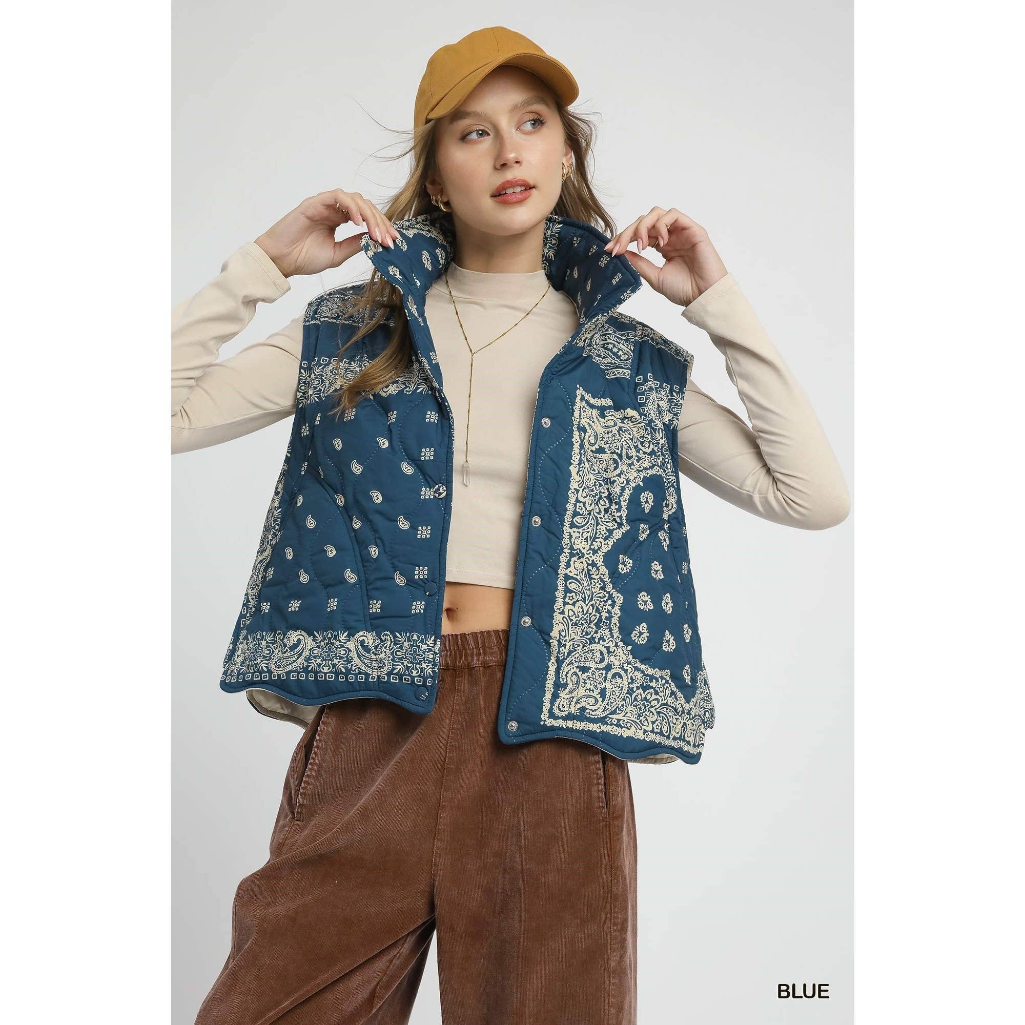 Woman wearing a blue patterned vest over a beige long-sleeve top and brown pants, with a mustard yellow cap.