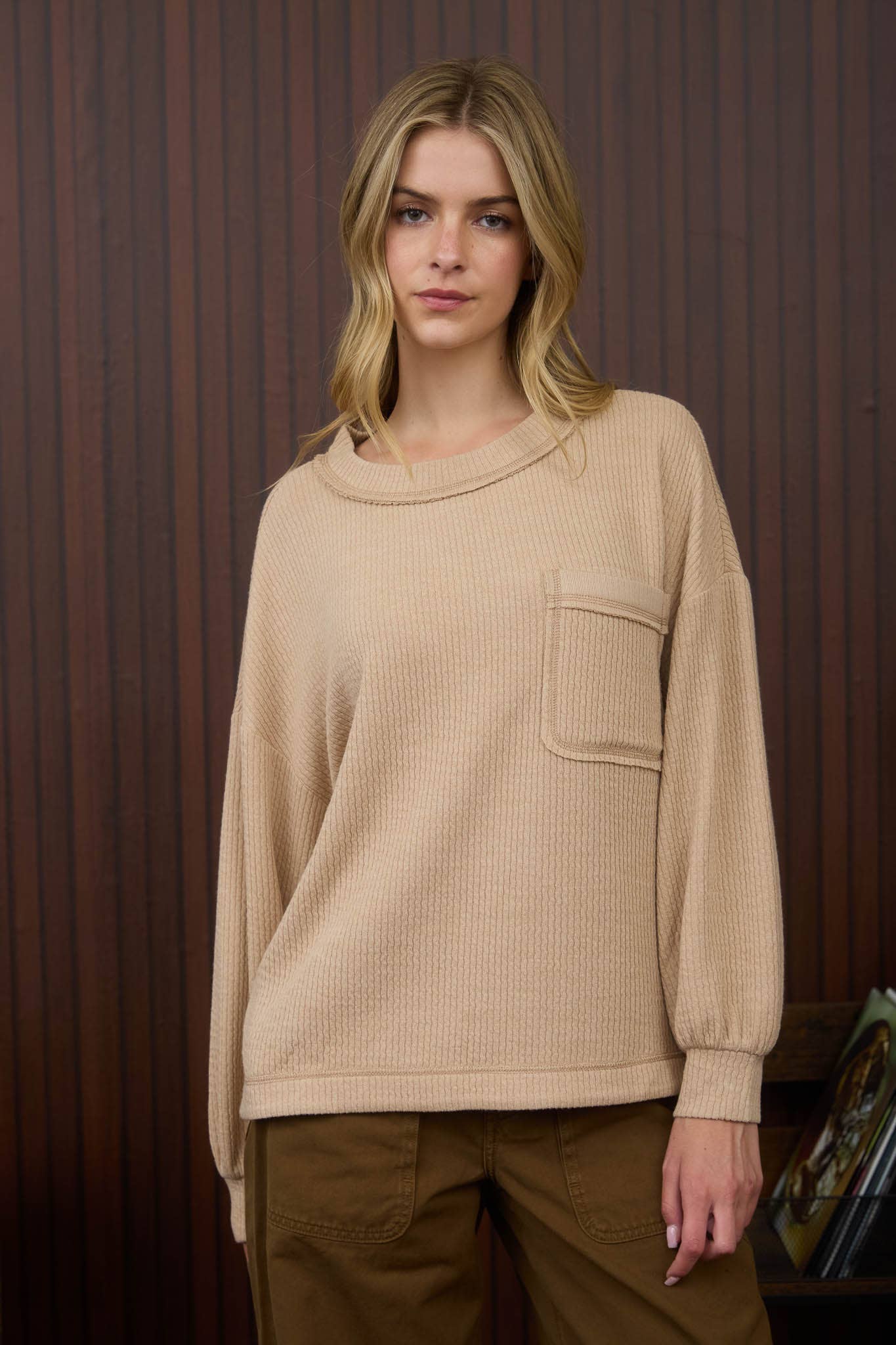Woman wearing a beige sweater with a pocket against a wooden paneled wall.