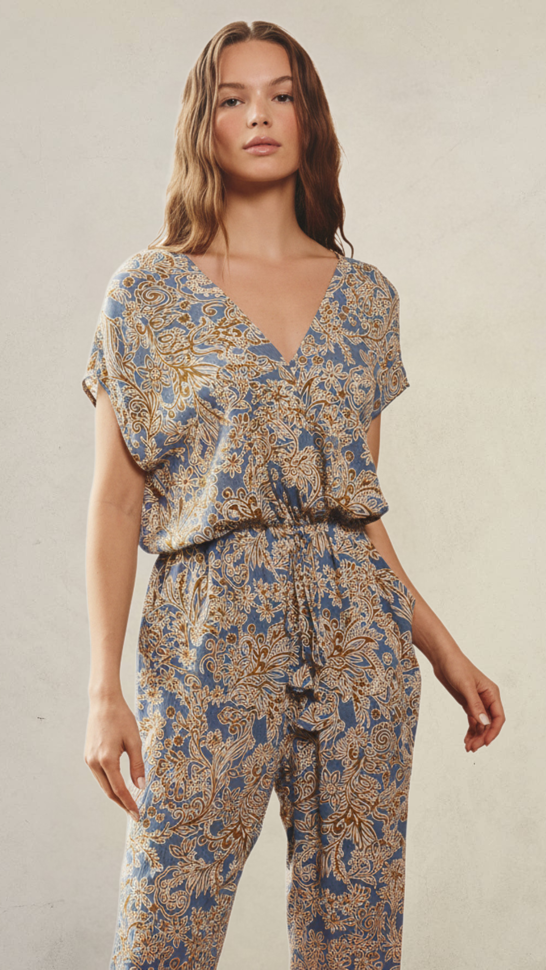 Model wearing the Blue Sage Jumpsuit, a soft blue paisley print one-piece featuring a surplice V-neckline, dolman sleeves, adjustable drawstring waist, side pockets, and tapered elastic ankle cuffs.