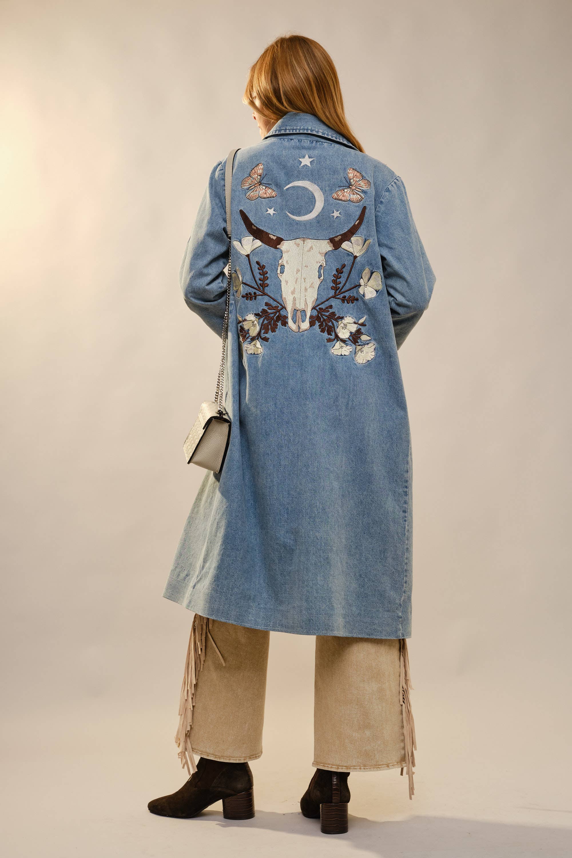 Person wearing a long denim coat with embroidered designs on a plain background