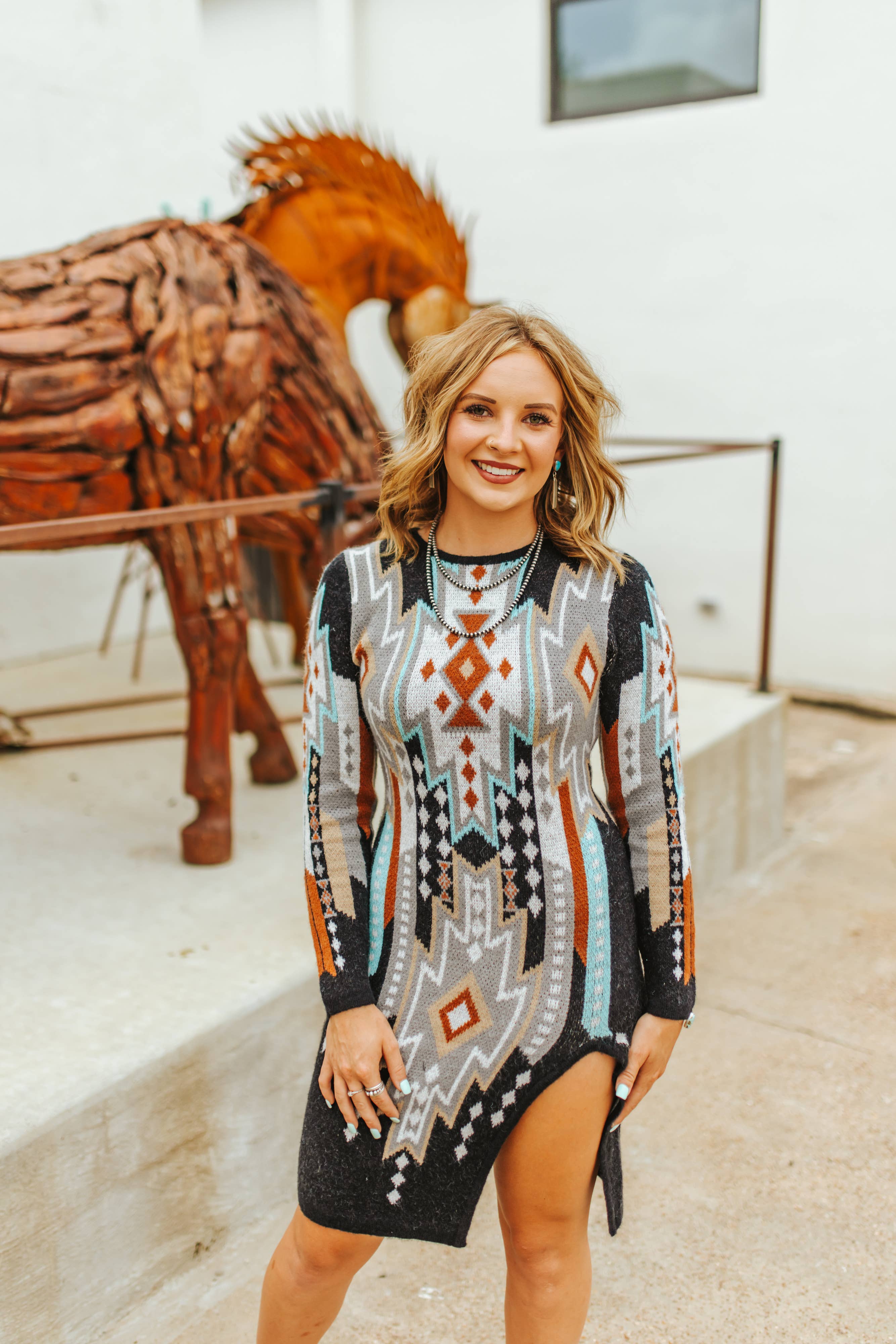 Woman wearing a patterned dress standing in front of a wooden horse sculpture.