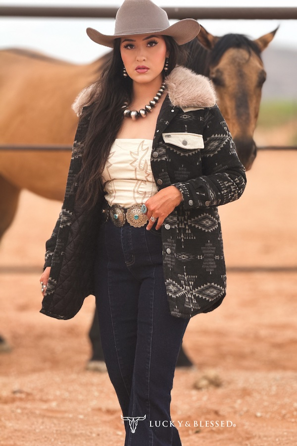 Woman in cowboy hat and patterned jacket standing next to a horse in an outdoor setting.