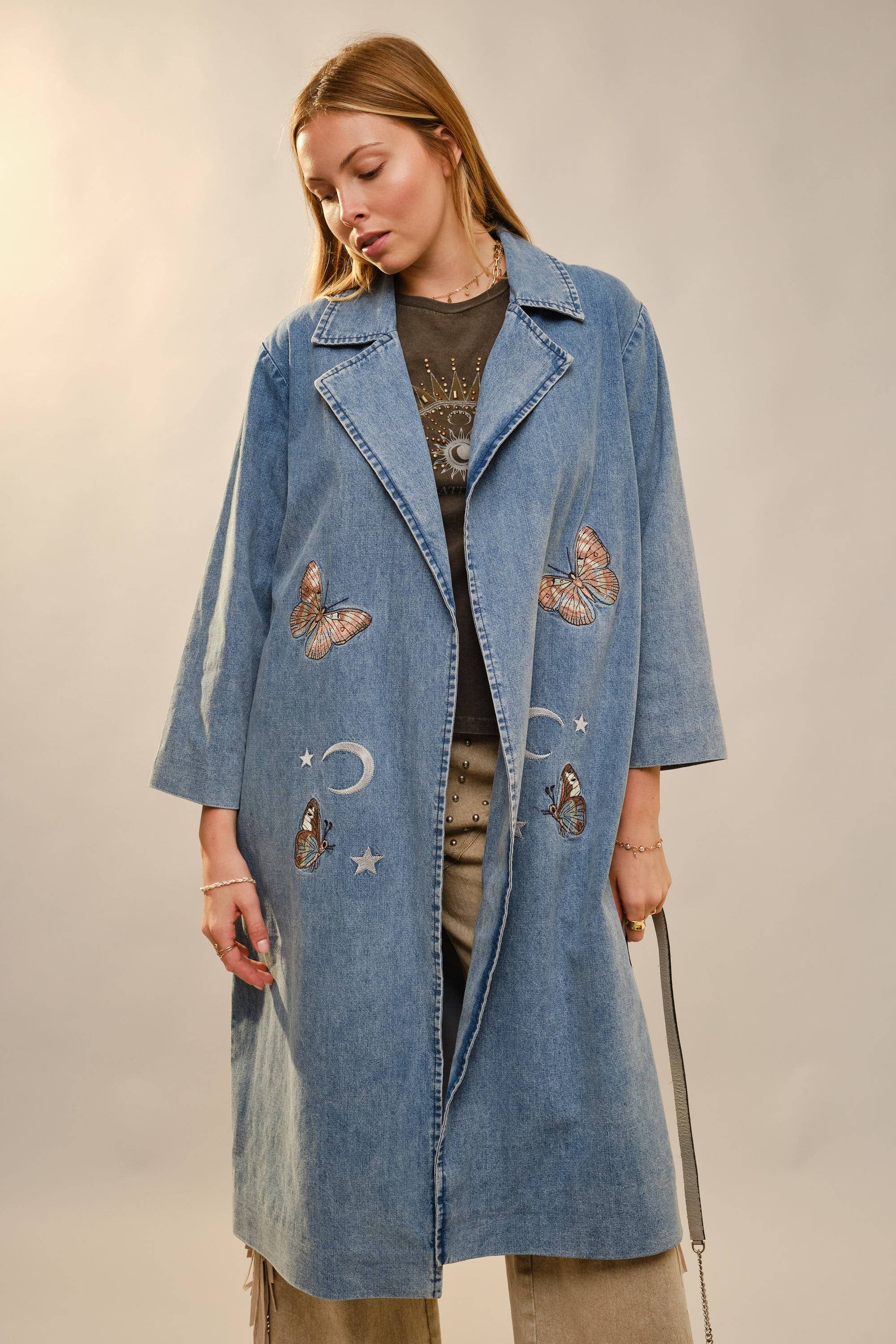 Woman wearing a long denim coat with embroidered designs on a neutral background