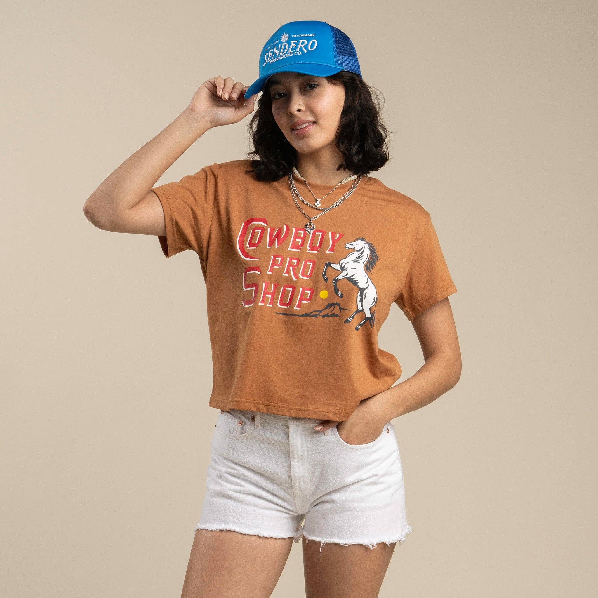Rust-colored cropped t-shirt featuring a Cowboy Pro Shop graphic with vintage Western lettering and a rearing horse design. Relaxed fit with a soft, lived-in feel.