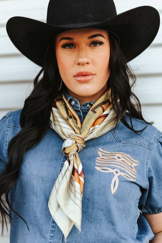 Woman wearing a black cowboy hat, denim shirt with embroidery, and patterned scarf against a white background.