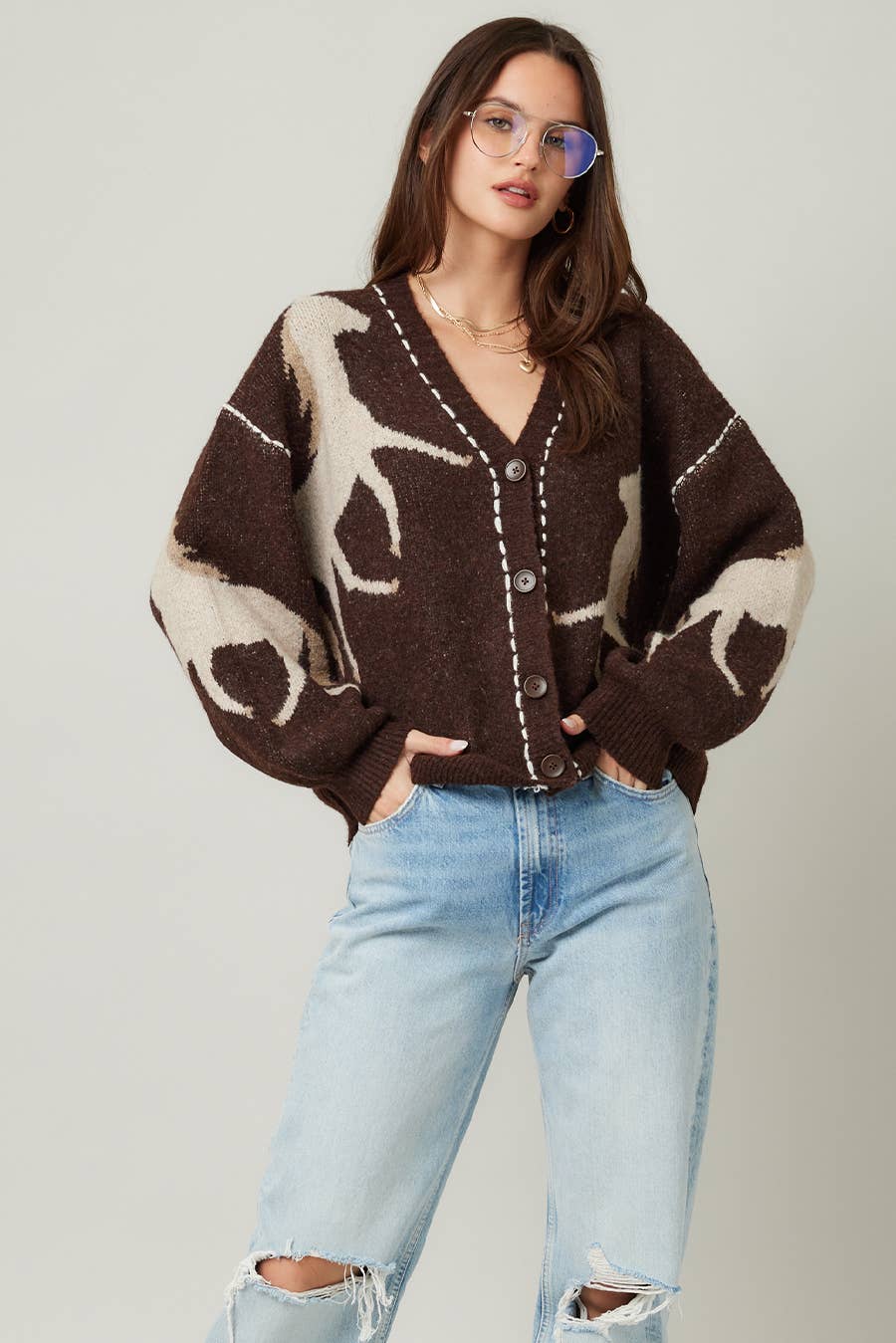 Woman wearing a brown cardigan with abstract patterns and light blue jeans on a plain background
