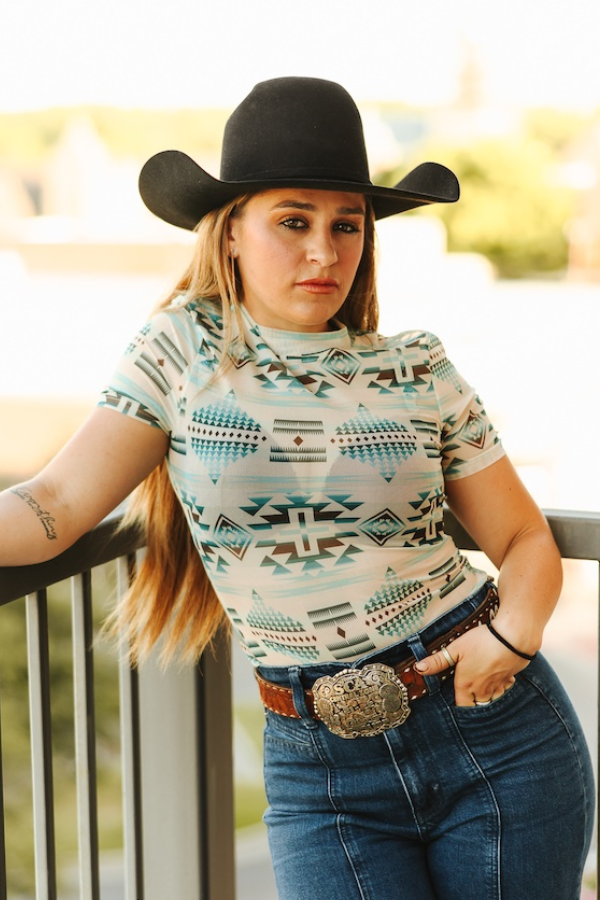 Woman wearing a patterned shirt, black cowboy hat, and blue jeans with a belt and large buckle.