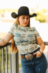 Woman wearing a patterned shirt, black cowboy hat, and blue jeans with a belt and large buckle.