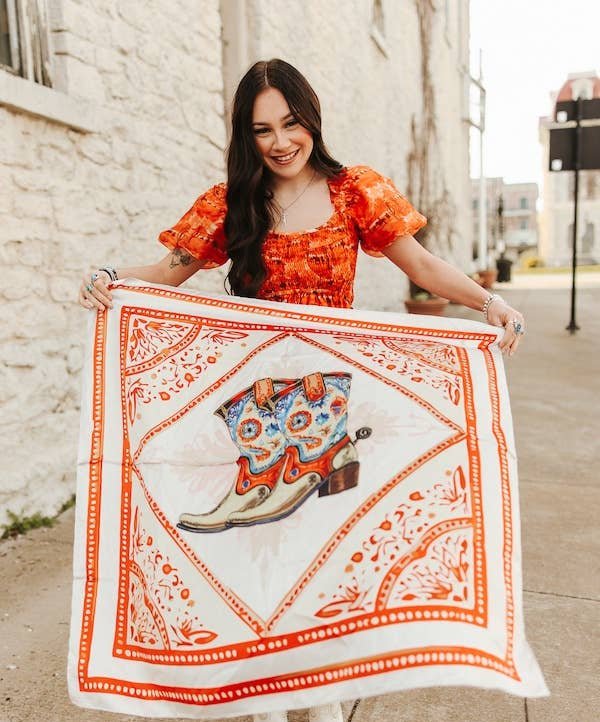 Woman holding a bandana with a cowboy boot design on a sidewalk.