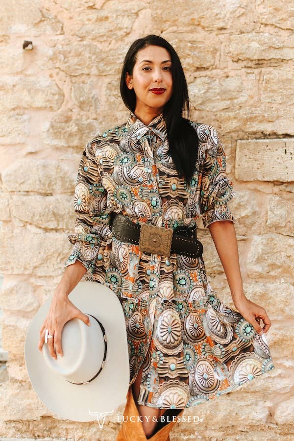 Woman wearing a patterned dress with a belt, holding a white hat against a stone wall.