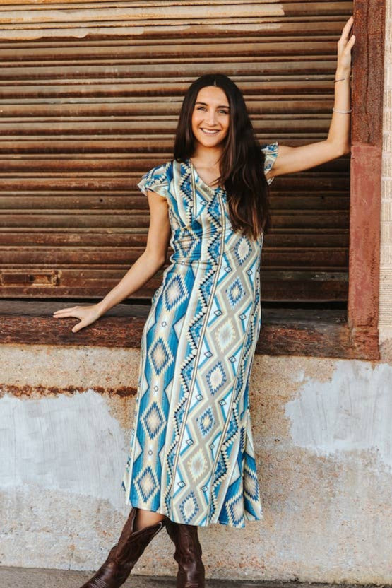 Woman in a patterned dress standing against a rustic wall.