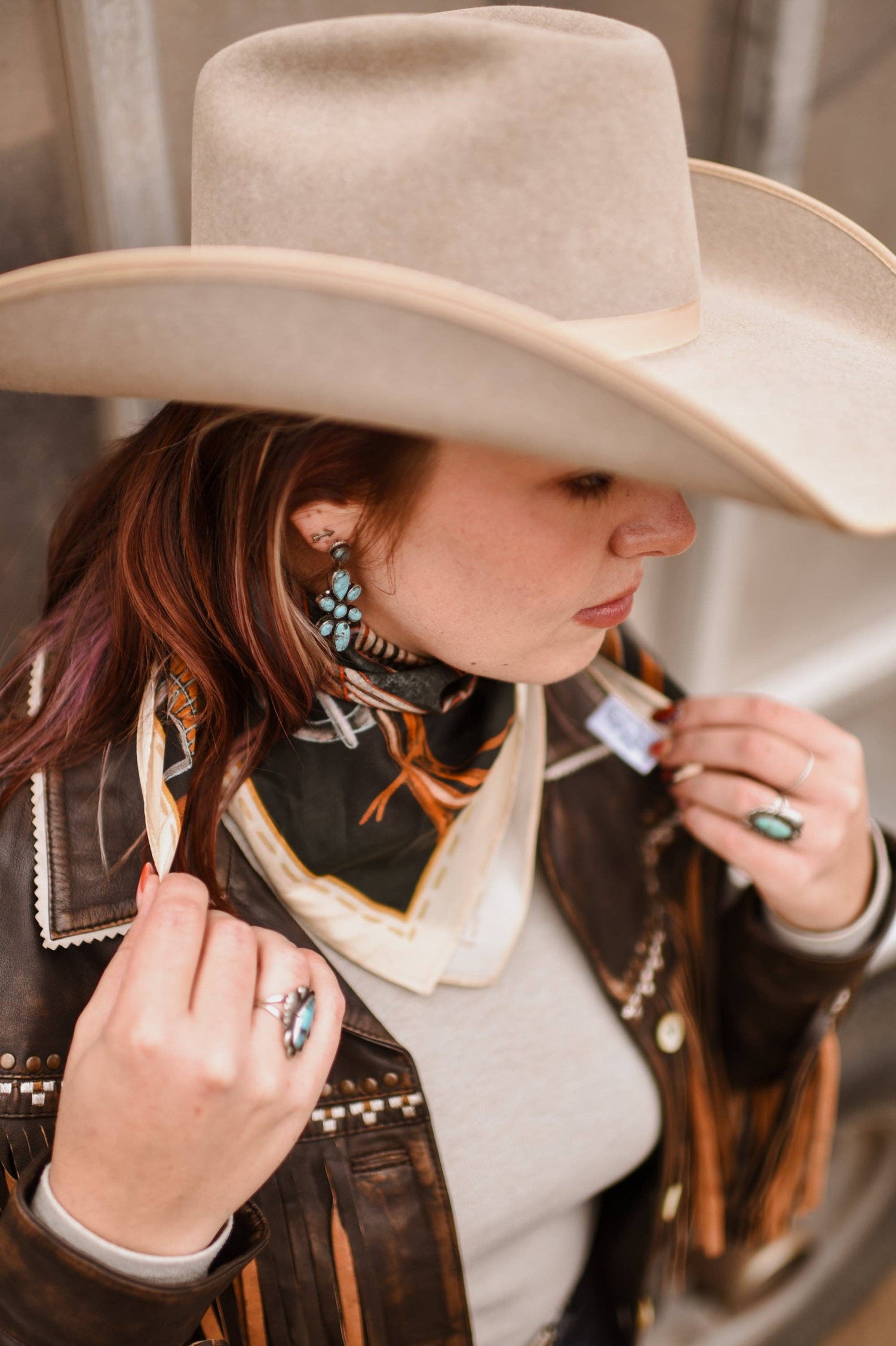 Person wearing a beige cowboy hat and brown leather jacket with a scarf, holding a small object.