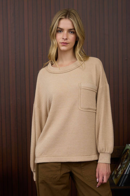 Woman wearing a beige sweater with a pocket against a wooden paneled wall.