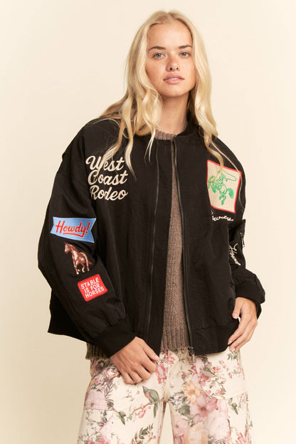 Woman wearing a black bomber jacket with patches on a beige background