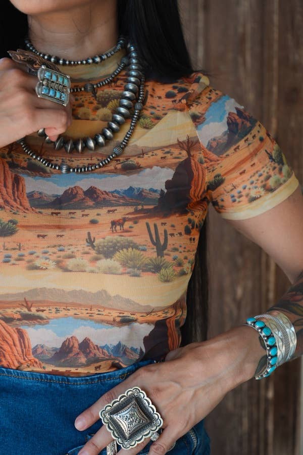 Person wearing a shirt with desert landscape print, holding a cup, with jewelry and tattoos visible.