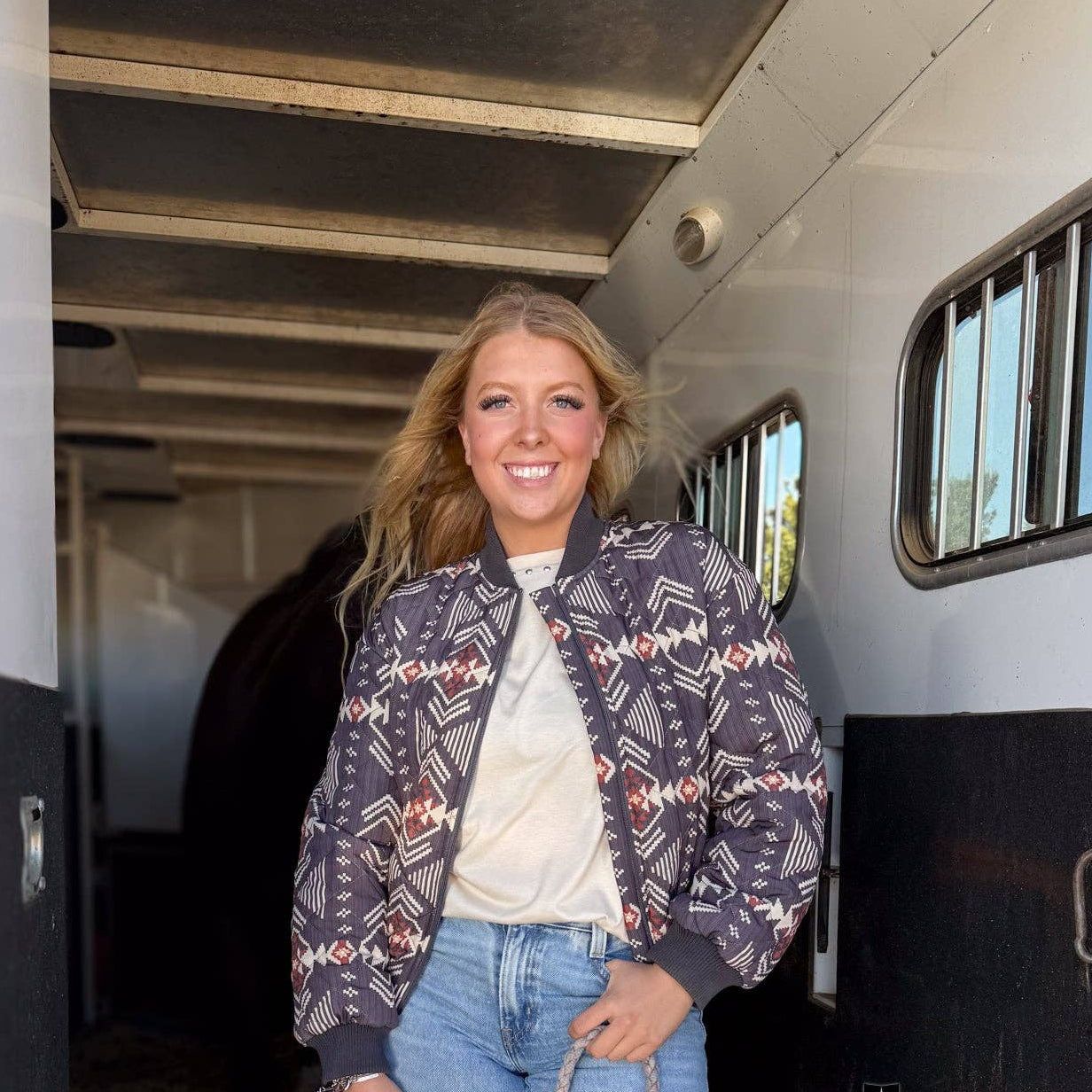 Woman standing next to a horse trailer holding a rope, wearing a patterned jacket and ripped jeans.