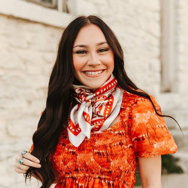Woman wearing an orange dress with a patterned scarf, standing against a light-colored wall.