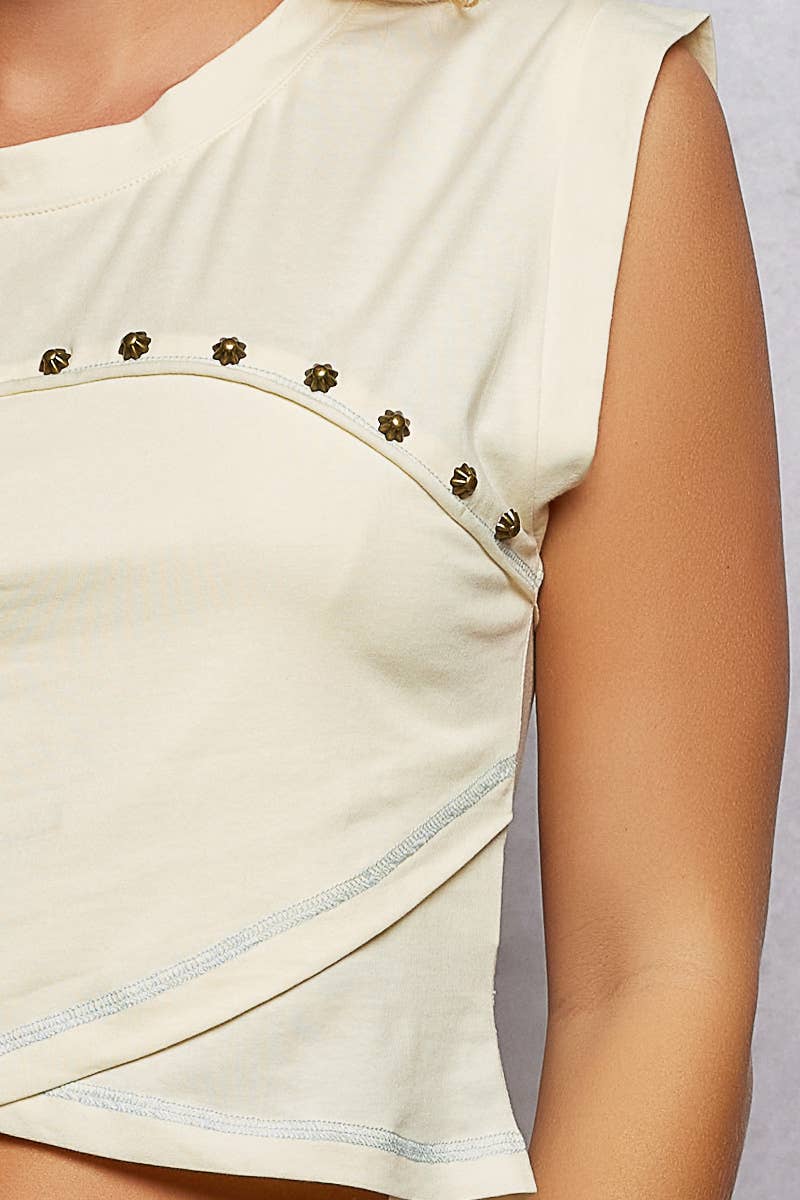 Model wearing the Ivory Trail Top, a cream cotton jersey sleeveless top featuring a round neckline, contrast exposed stitching, stud detailing across the chest, an asymmetrical wrap-style high-low hem