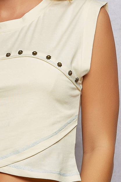 Model wearing the Ivory Trail Top, a cream cotton jersey sleeveless top featuring a round neckline, contrast exposed stitching, stud detailing across the chest, an asymmetrical wrap-style high-low hem