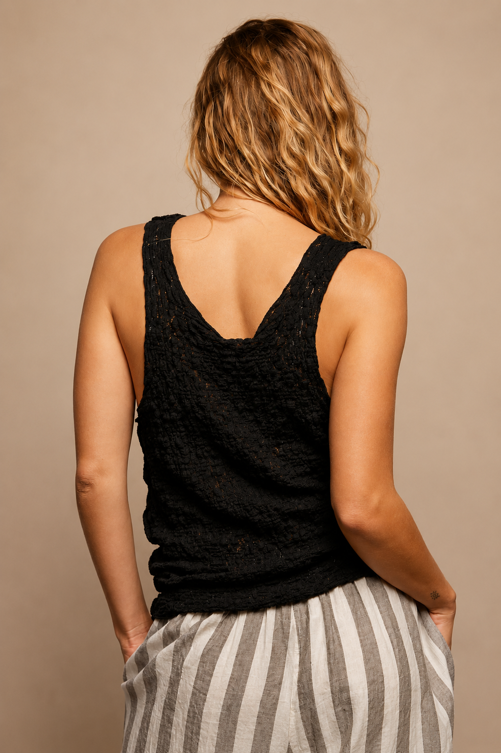 Model wearing the Midnight Lace Tank, a black sleeveless scoop-neck top featuring intricate floral lace panels, textured fabric, exposed seam detailing, and a relaxed fit, paired with striped linen pants.