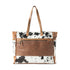 S-10917 Cowhide Tooled Leather Tote featuring a genuine hair-on hide body, intricate floral tooled leather panel, sturdy leather handles with rivet details, and a spacious design with exterior and interior pockets for functionality and style.