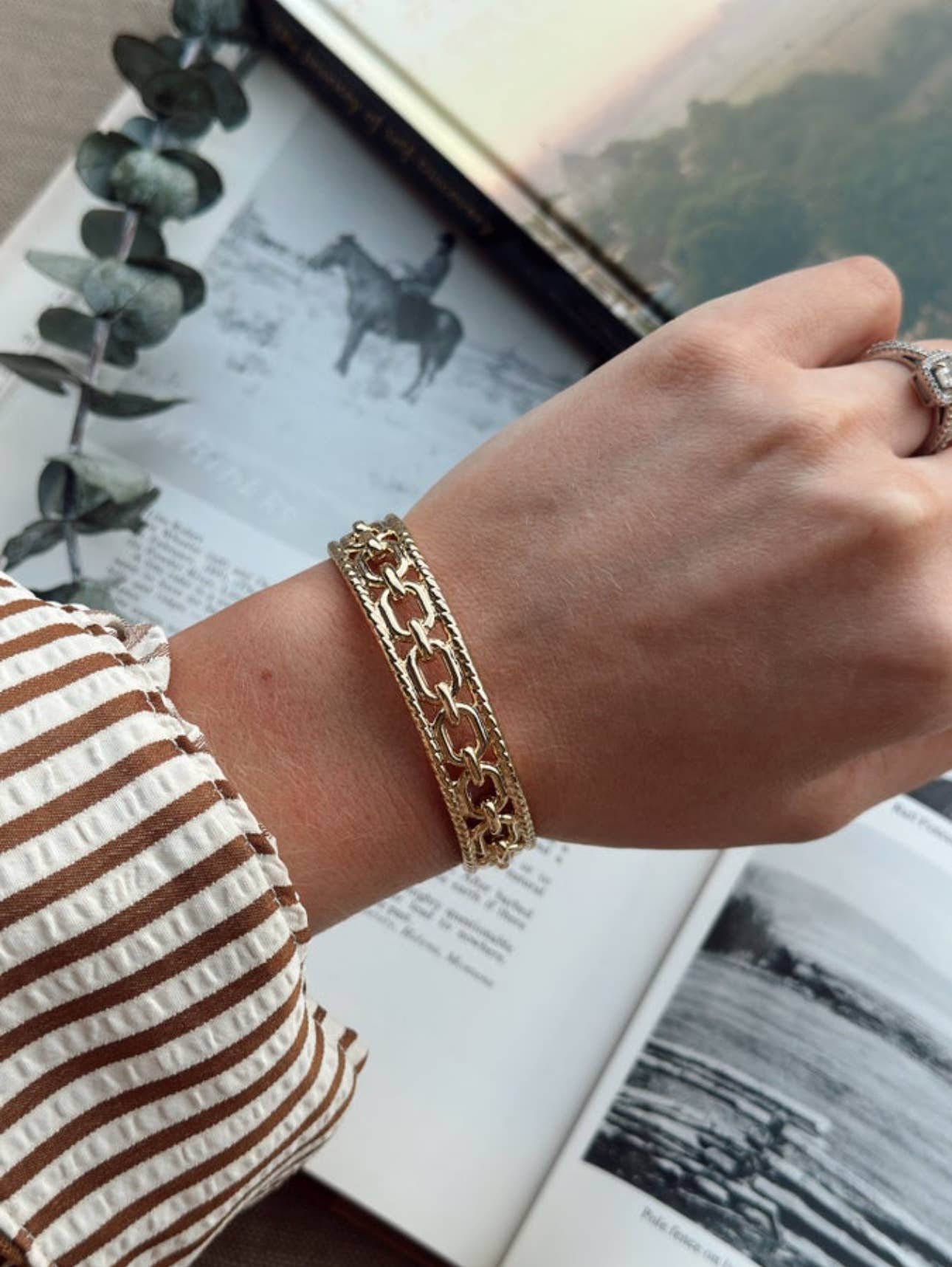 Gold chain bracelet on a wrist with an open book and eucalyptus leaves in the background