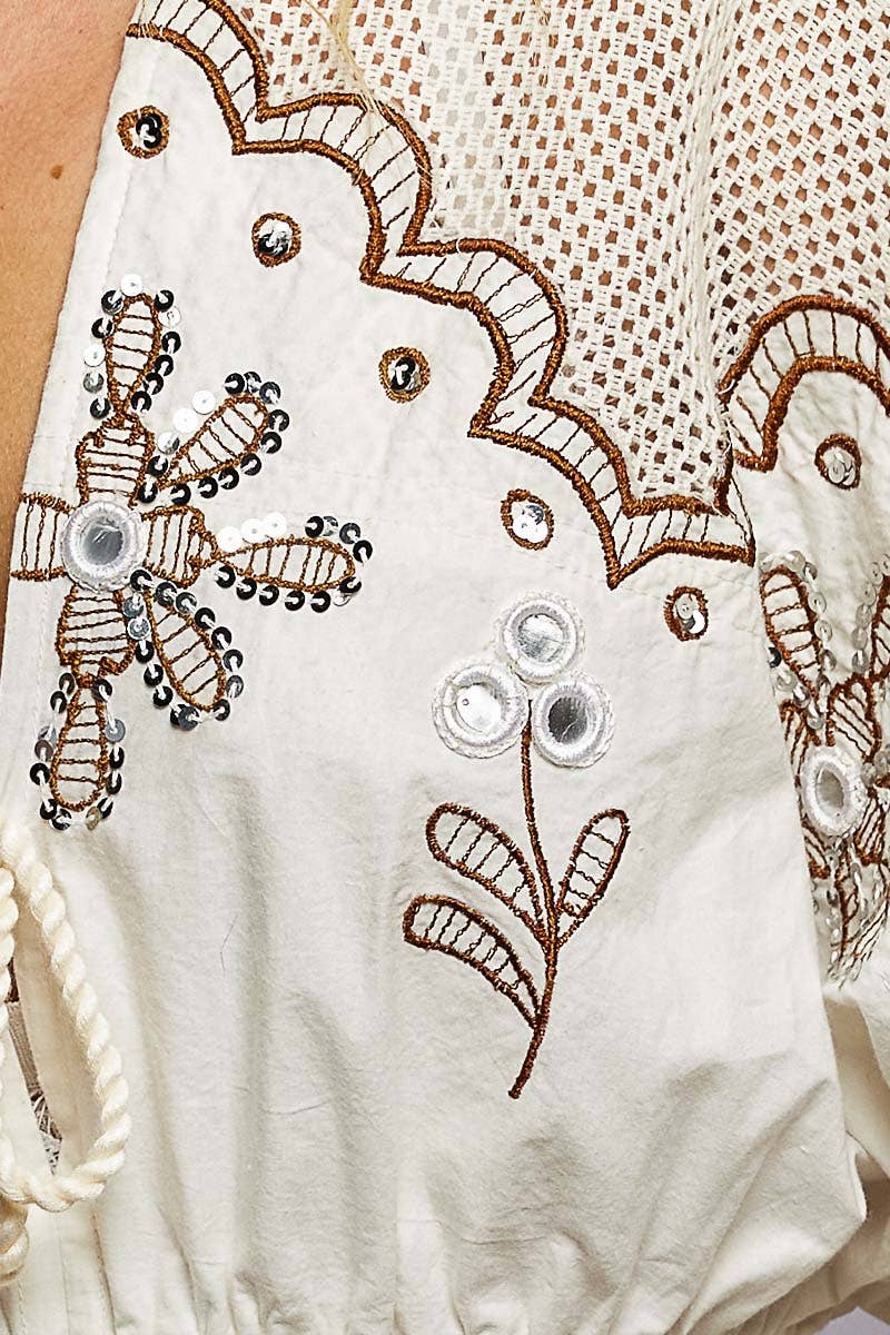 Model wearing The Marfa Blouse, a cream cropped cotton top featuring embroidered detailing, mirror accents, crochet shoulder panels, tasseled ties, flutter sleeves, an elastic waist, and a ruffled hem, paired with pink striped pants.