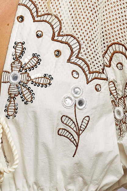 Model wearing The Marfa Blouse, a cream cropped cotton top featuring embroidered detailing, mirror accents, crochet shoulder panels, tasseled ties, flutter sleeves, an elastic waist, and a ruffled hem, paired with pink striped pants.