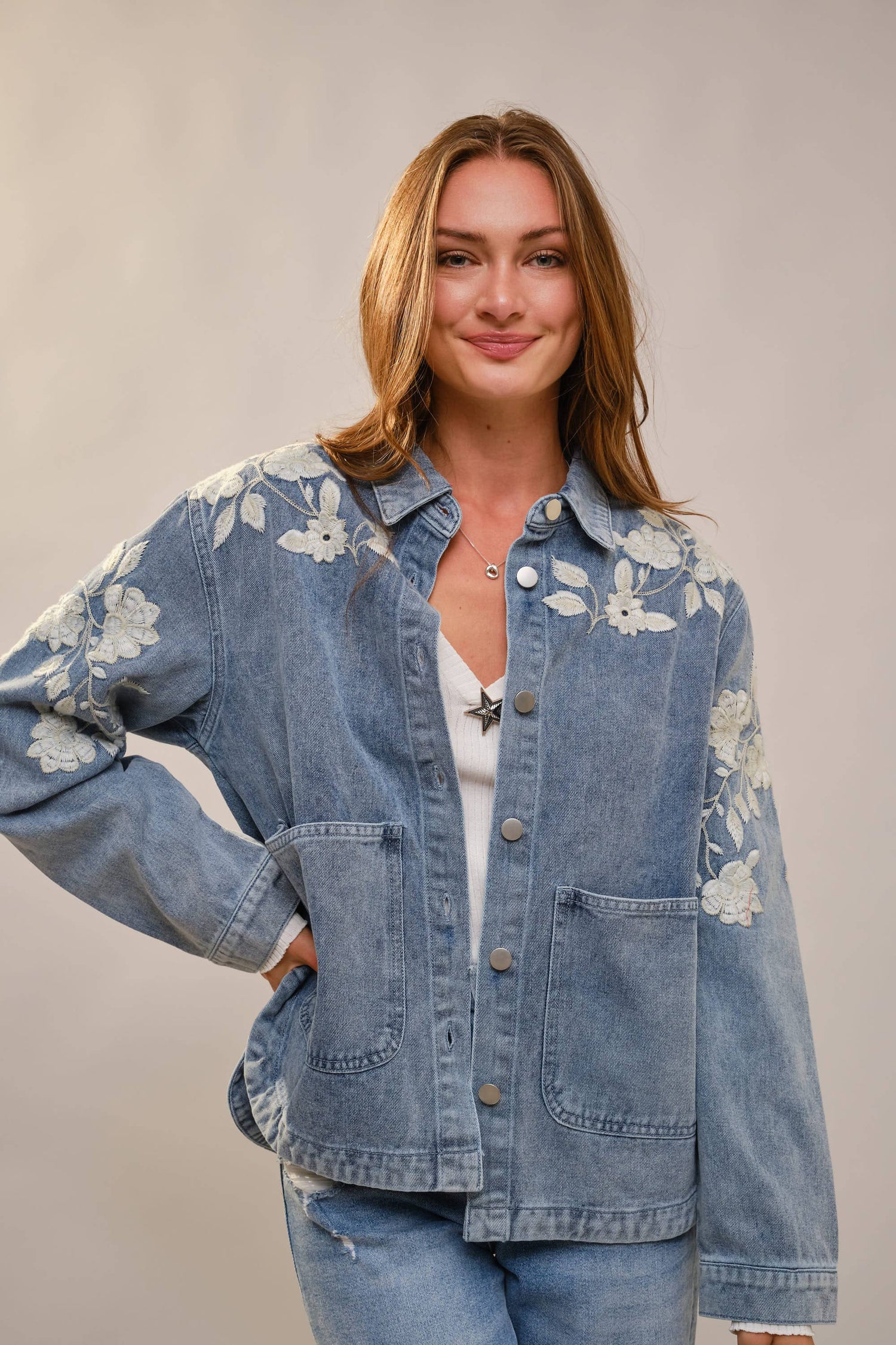 Woman wearing a blue denim jacket with floral embroidery on a plain background