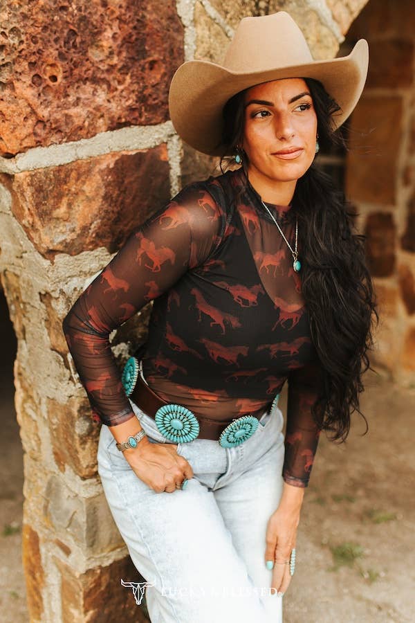 Woman wearing a cowboy hat and patterned top leaning against a stone wall.
