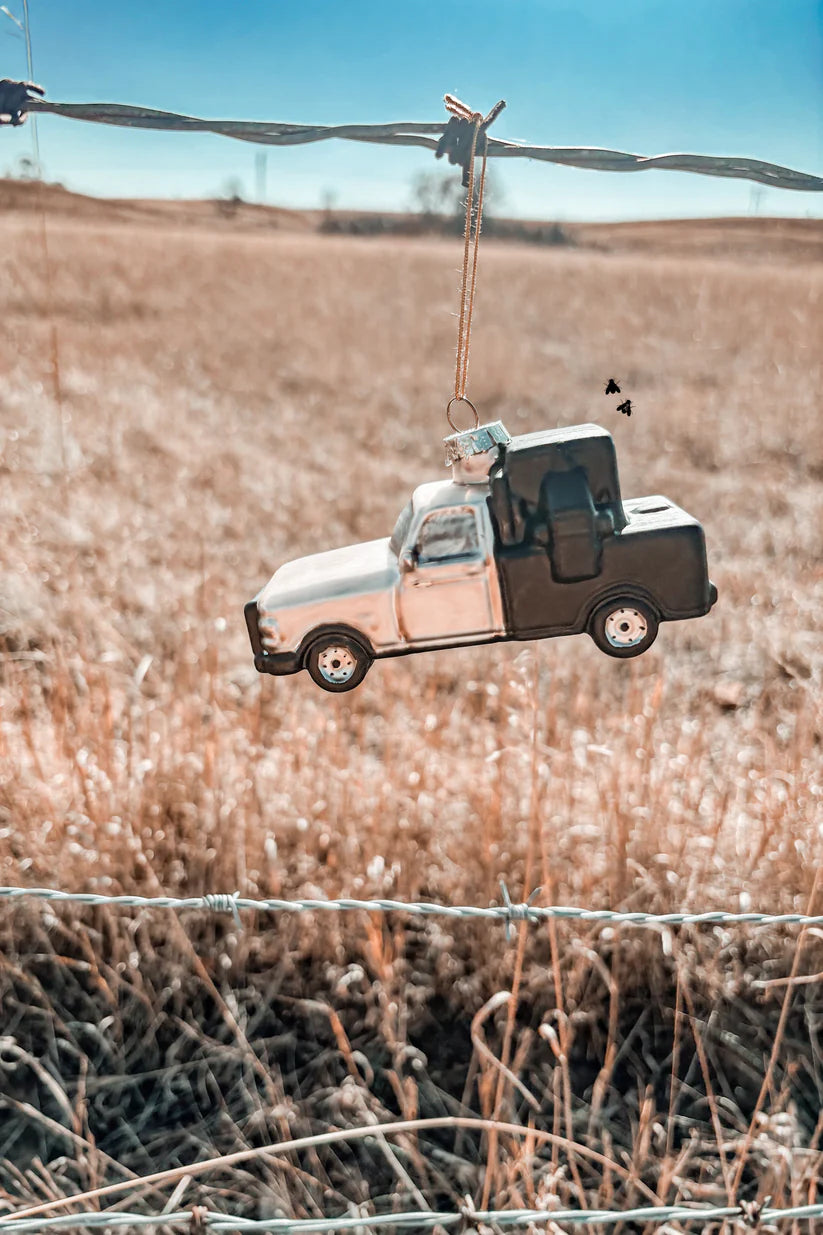 A blown glass ornament in the shape of a classic truck, hanging from a wire in a field with a blue sky in the background.