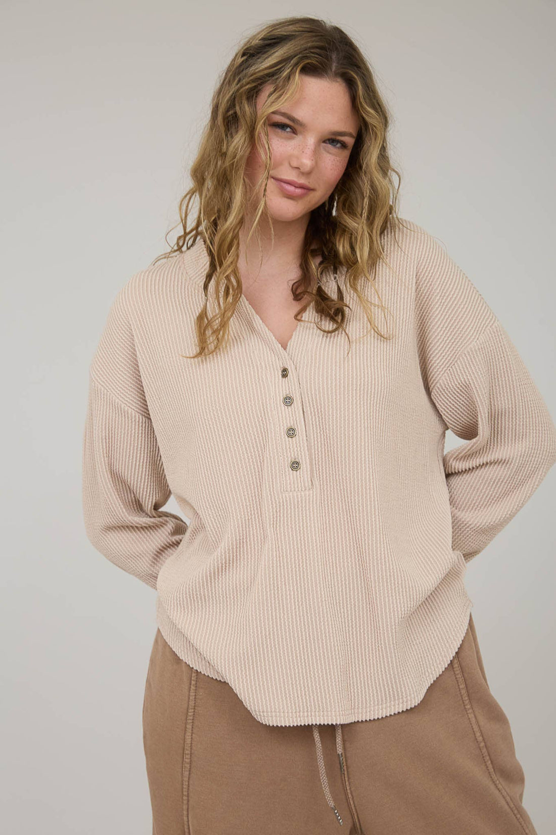 Woman wearing a beige ribbed long-sleeve shirt and brown pants against a plain background