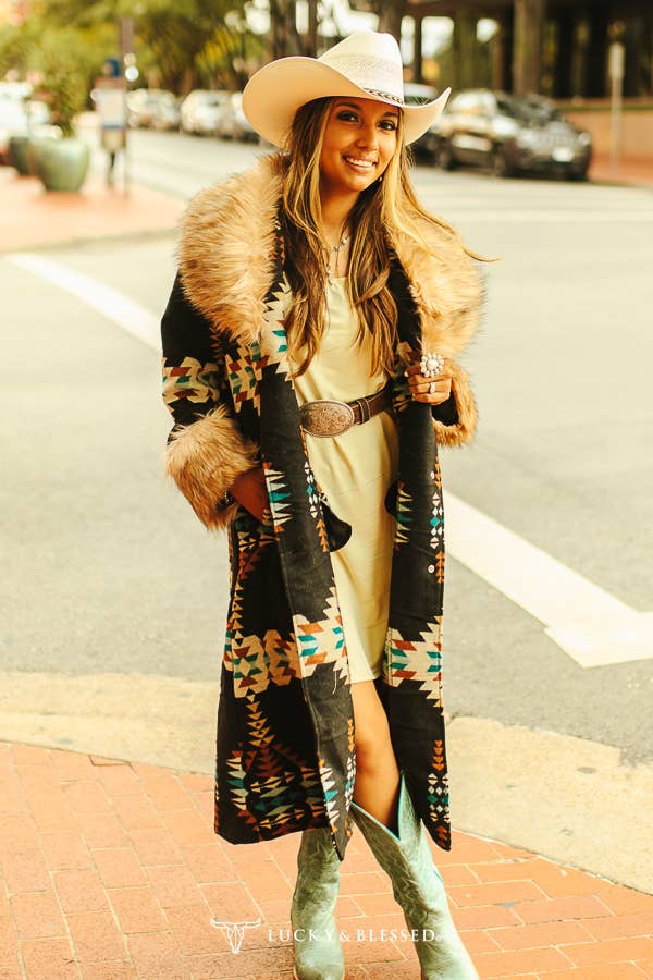 Woman wearing a patterned coat with fur trim and a cowboy hat on a street.