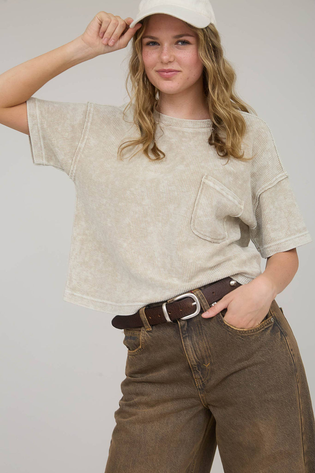 Person wearing a beige oversized shirt, brown pants, and a white cap on a plain background